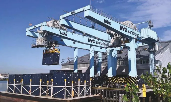Large blue waterfront cranes transfer shipping containers between a terminal and barges.
