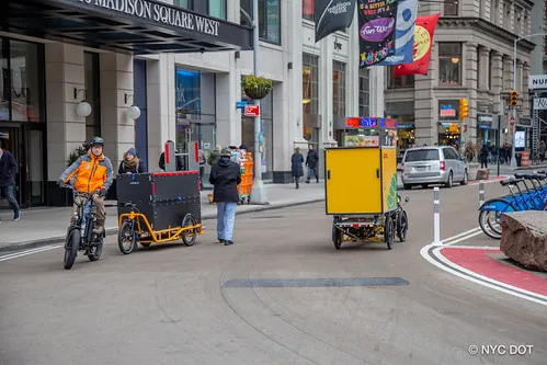 Two commercial cargo bikes travel along a New York City street, including a yellow DHL cargo bike.